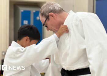 Tony is pointing at a child's feet and giving instructions to the youngster. Tony has grey hair and is wearing glasses. He has a black belt around his white judo outfit. The child has black hair and is also wearing a white judo strip