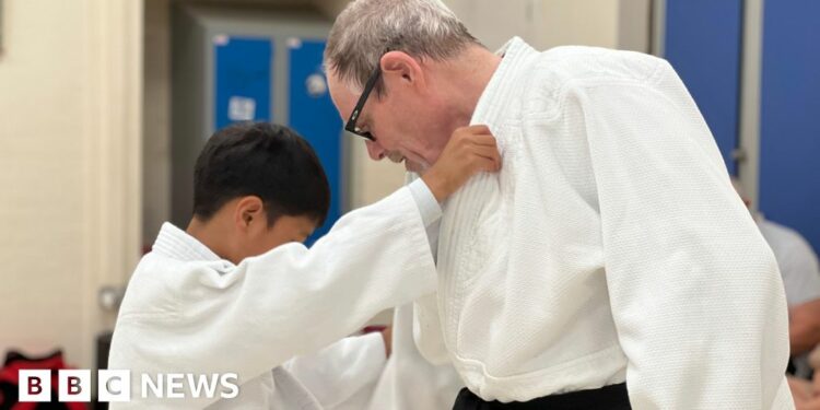 Tony is pointing at a child's feet and giving instructions to the youngster. Tony has grey hair and is wearing glasses. He has a black belt around his white judo outfit. The child has black hair and is also wearing a white judo strip