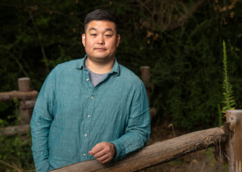 Taeyoon Lee, Ph.D. He is standing with his arm on a wooden rail. He is wearing a green button down shirt