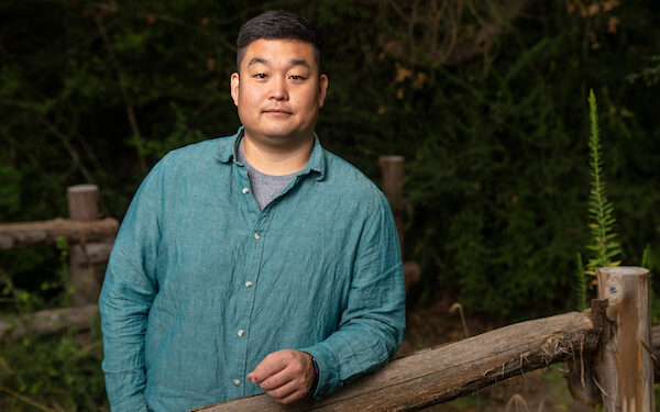 Taeyoon Lee, Ph.D. He is standing with his arm on a wooden rail. He is wearing a green button down shirt