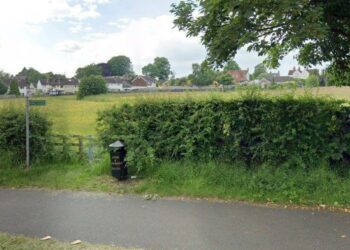 A public footpath sign pointing across a grassy field next to a metal gate and a black litter bin next to it. Houses are in the distance.