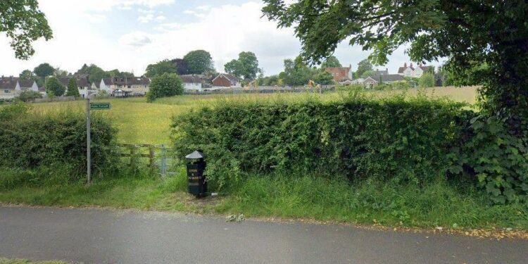 A public footpath sign pointing across a grassy field next to a metal gate and a black litter bin next to it. Houses are in the distance.