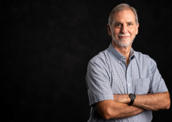 A man stands with his arms folded. He s wearing a blue checkered short and he has a gray beard.