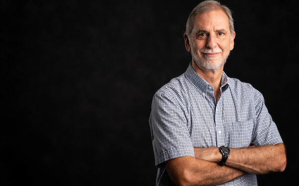 A man stands with his arms folded. He s wearing a blue checkered short and he has a gray beard.